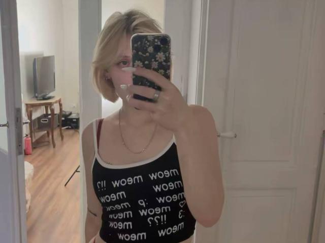 AnnaCandys with short blonde hair, wearing a black tank top showing "warm" text, holding a phone with a floral case, taking a mirror selfie, showing her tits.
