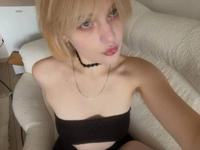 AnnaCandys with blonde hair, pouty lips, black choker, and black strapless top, sitting on a white couch, showing her bare shoulders and small tits.