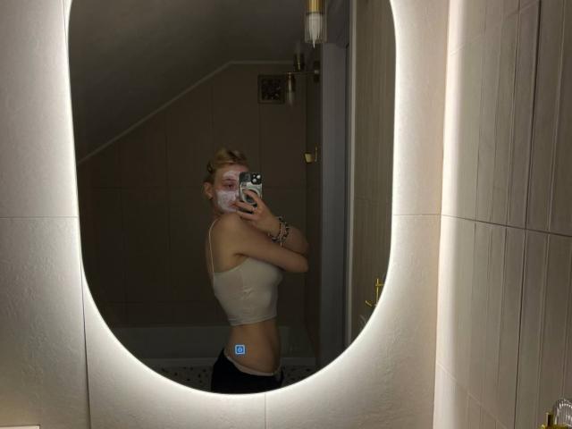 AnnaCandys, blonde, in a beige tank top, black pants, holding phone, mirror selfie, face covered in white paste, bathroom, oval mirror, soft lighting, visible butt, slim.