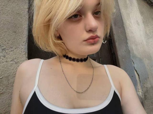 AnnaCandys with blonde hair, wearing a black choker, chain necklace, and black-white tank top, showing small tits, pale skin, and a serious expression, standing in front of a wall.