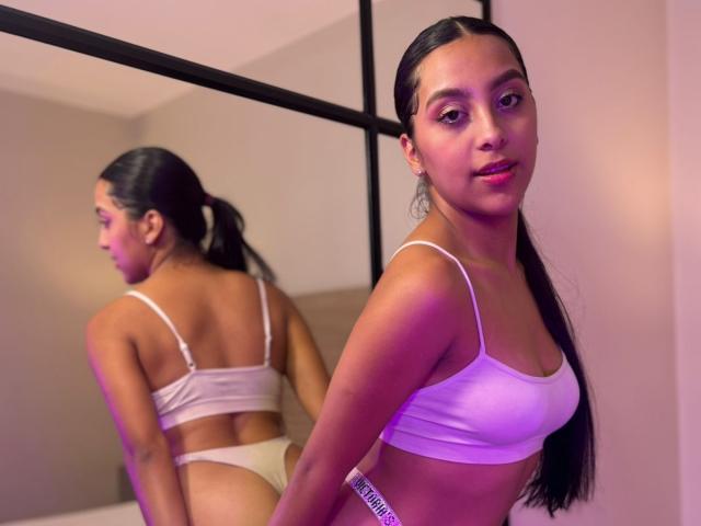 EmmaEstreilla with long black hair, white bra, and white panties, standing in front of a mirror, showing her back and ass, looking at the camera, smiling.