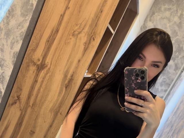 AdelaideLisa, avec de longs cheveux noirs, un haut noir sans manches, tenant un téléphone avec une coque à motifs floraux, prenant un selfie devant un miroir, montrant ses seins, une porte en bois, des murs gris texturés, et une bague à sa main droite..
