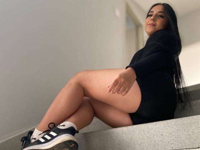 StefanyDelly with long black hair, black sweater, and black shorts, sitting on gray stairs, legs crossed, wearing black and white Adidas sneakers, smiling slightly.