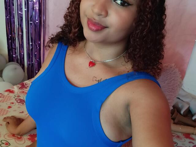 JessyZoee with curly brown hair, blue tank top, red heart necklace, smiling, pink lipstick, medium tits, light brown skin, and a cute face, holding a stuffed animal.
