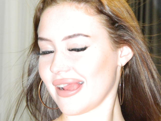 MicheleJinkins has her tongue out, showing a small silver piercing, with eyes closed, wearing long, dark brown hair and large gold hoop earrings. Bright, white skin.