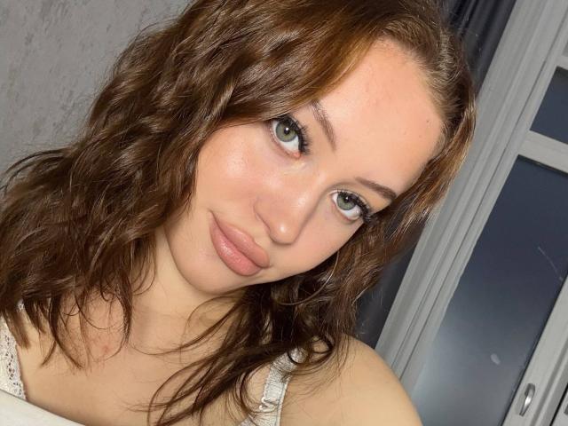 MicheleJinkins with wavy brown hair, green eyes, and pink lips, wearing a white lace top, looking at the camera with a subtle smile. Her ass is not visible.