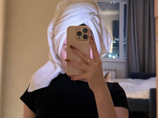 MicheleJinkins with a white towel wrapped around her head, black shirt, holding a pink phone, covering her face, red nails, in a dimly lit room, city lights visible.