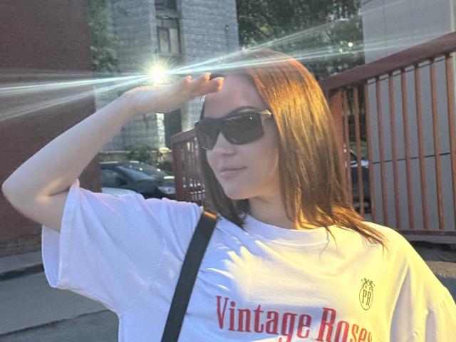 MicheleJinkins with brown hair, wearing black sunglasses, white "Vintage Road" shirt, raising hand to shield eyes from sun, black shoulder strap, street in background.