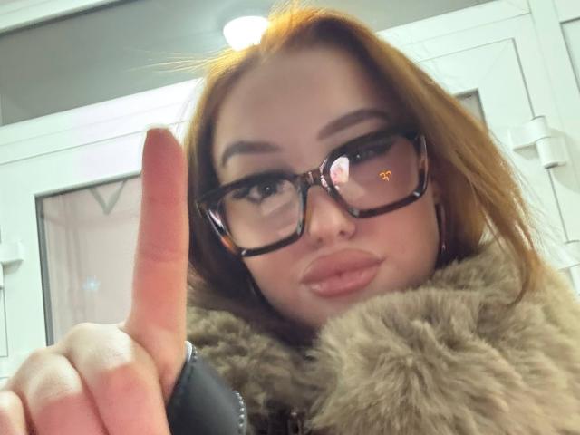 MicheleJinkins with glasses, red hair, and pouty lips, wearing a fur coat, giving a middle finger with her right hand, reflection of a light in her glasses.