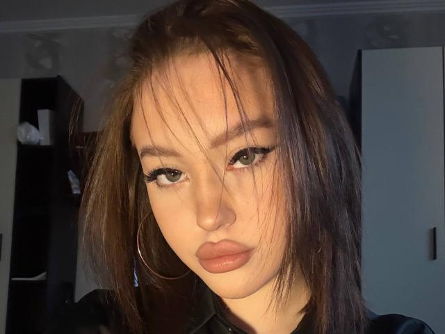 MicheleJinkins has dirty brown hair, green eyes, and full lips with a serious look, wearing large hoop earrings and a black shirt, illuminated by warm indoor light.