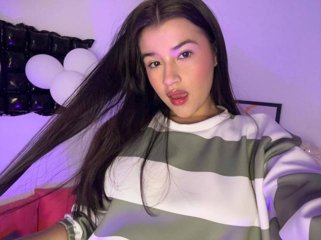 IrisBlair with long black hair, green and white striped shirt, sticking out tongue, purple eyeshadow, fair skin, purple light, white balloons in background.