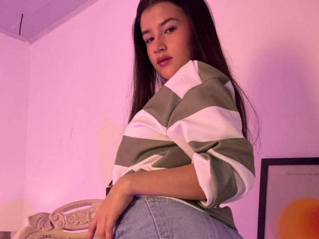 Iris Blair, met lang donker haar, een groen-wit gestreepte blouse en blauwe jeans, kijkt naar beneden met een lichte grijns, haar handen op haar billen. In de achtergrond is een rozeachtige kamerlicht te zien..