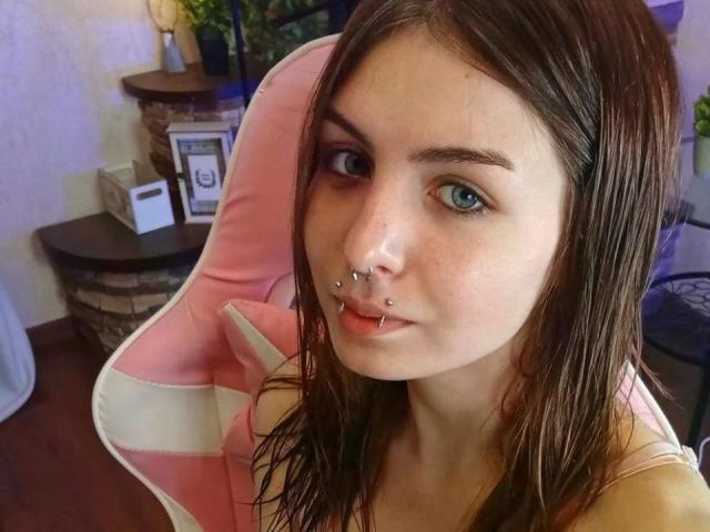NobukoGraue with wet brown hair, blue eyes, nose piercings, and a lip piercing, sitting in a pink-and-white chair, looking at the camera. Boobs not visible.