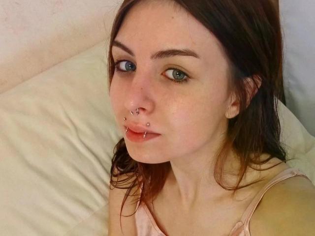 NobukoGraue has fair skin, green eyes, brown hair, and nose piercings with lip piercings. She's wearing a pink top, and her small tits are barely visible.
