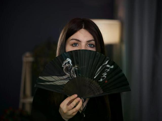 NobukoGraue holding a dark green fan with a white dragon pattern, partially covering her blue eyes. She has long brown hair and is wearing a black top.