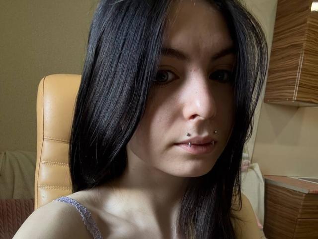NobukoGraue with black hair, nose piercing, and lip piercing, wearing a purple lace top, sitting in a tan leather chair, looking slightly annoyed.