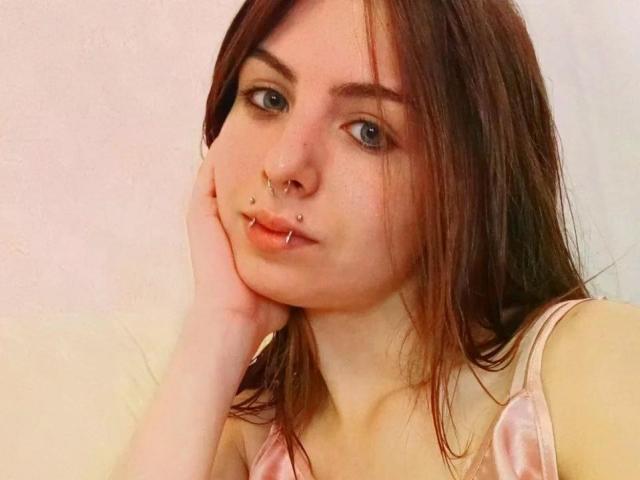 NobukoGraue has brown hair, blue eyes, and multiple facial piercings, including a nose ring and lip studs. She's wearing a pink tank top, resting her cheek on her hand.