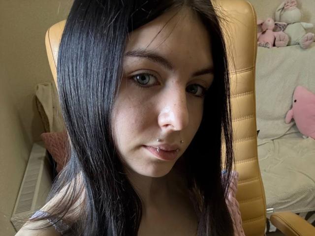 NobukoGraue has pale skin, dark straight hair, green eyes, and a lip piercing. She's sitting in a yellow leather chair, showing a slight smile. Her tits are small.