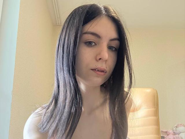 NobukoGraue has straight brown hair, green eyes, a nose piercing, and a lip piercing, with a light skin tone, sitting in a chair, wearing a strapless top.