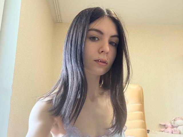 NobukoGraue with straight brown hair, light skin, and a septum piercing, wearing a lace top, looking at the camera with a slight open-mouth expression.