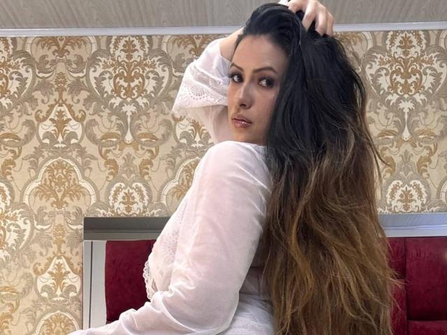 HannaRobertss with long, wavy brown hair, wearing a sheer white lace top, looking over her shoulder, hand in hair, red velvet couch, patterned wallpaper.