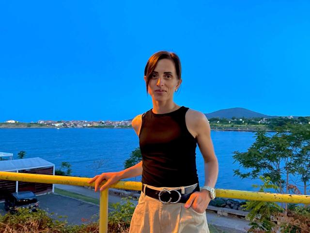 MilediElla-hot stands with hands in pockets, wearing a black sleeveless top, beige pants, and a belt with a circular buckle. She's leaning on a yellow railing, blue sky.