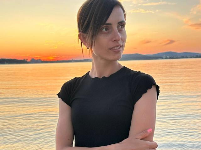MilediElla-hot with brown hair, small tits, black shirt, crossed arms, pink nails, smiling, standing by water at sunset, orange sky, mountains in background.