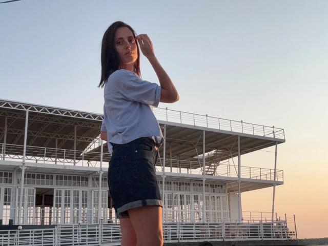 MilediElla-hot with medium tits, wearing a loose gray shirt and black denim shorts, standing in front of a white, metal-roofed structure at sunset, hand in hair.