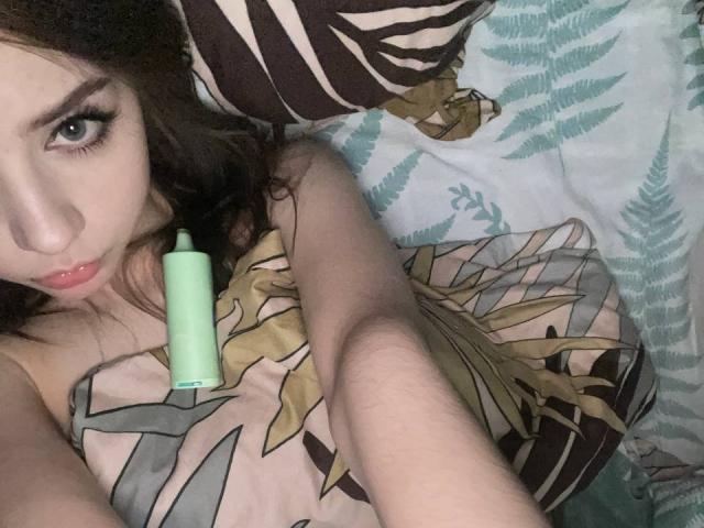 MorganCastellanoz com um vibrador verde no peito, deitada na cama com travesseiros de padrão de zebra e folha, olhando para cima com lábios curvados, cabelo cacheado escuro, pele clara e olhos azuis..