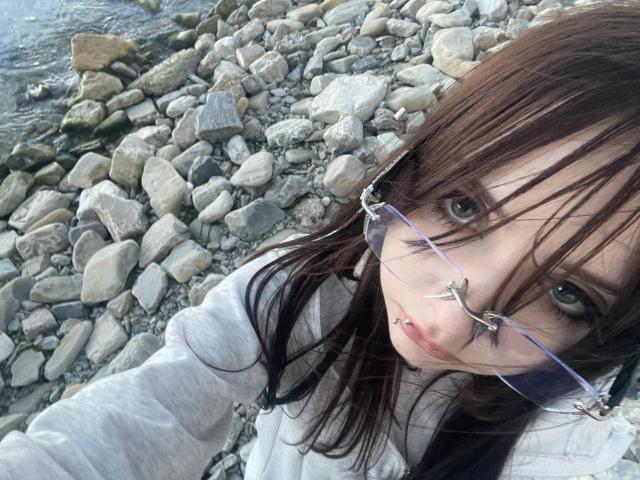 MorganCastellanoz with brown hair, glasses, nose ring, and lip piercing, wearing a gray hoodie, standing by a rocky riverbank.