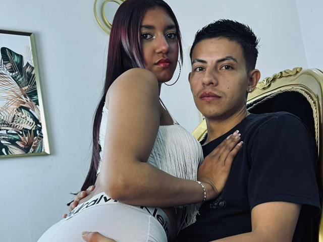 CelesteAndBlake with long red hair and white fringed top, hugging Blake's black shirt, showing her white pants and her ass. Blake has short black hair, looking at camera.
