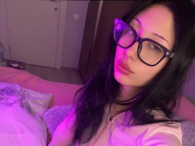 GlitchDoll with purple-lit face, black glasses, nose ring, and long black hair, showing off her pouty lips and subtle cleavage in a pink and purple room.