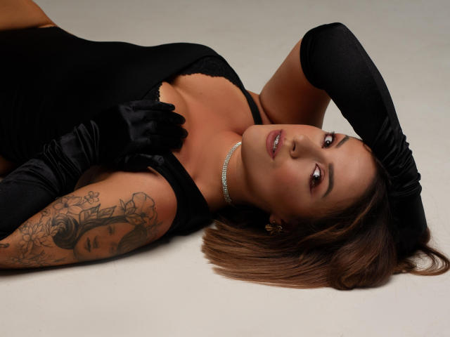 Molly, com luvas pretas, deitada de costas, mostrando o braço tatuado, vestindo um vestido preto, um colar brilhante e olhando para cima com um sorriso sedutor, com o cabelo castanho espalhado..