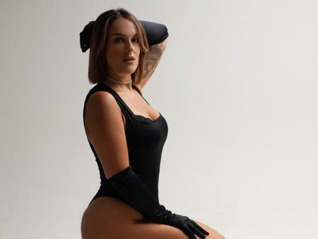 MollyPretty in black lingerie, sitting with arms behind head, wearing black gloves, showing her tits and ass, light brown hair, tattoo on right arm, white background.