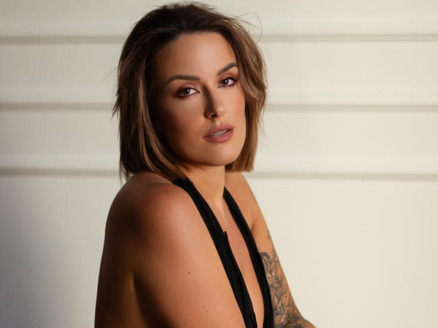MollyPretty has medium brown hair, brown eyes, and a tattoo on her right arm. She's wearing a black halter top, showing her bare shoulders and small tits.