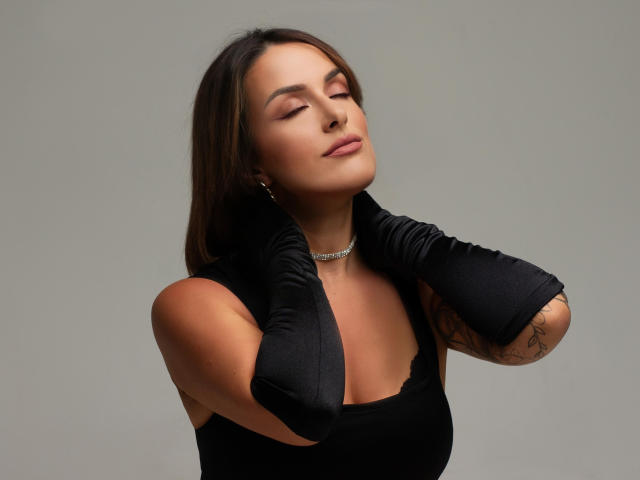 MollyPretty with eyes closed, wearing black gloves, choker, and a black top, touching her neck with a slight smile, showing a tattoo on her right arm.