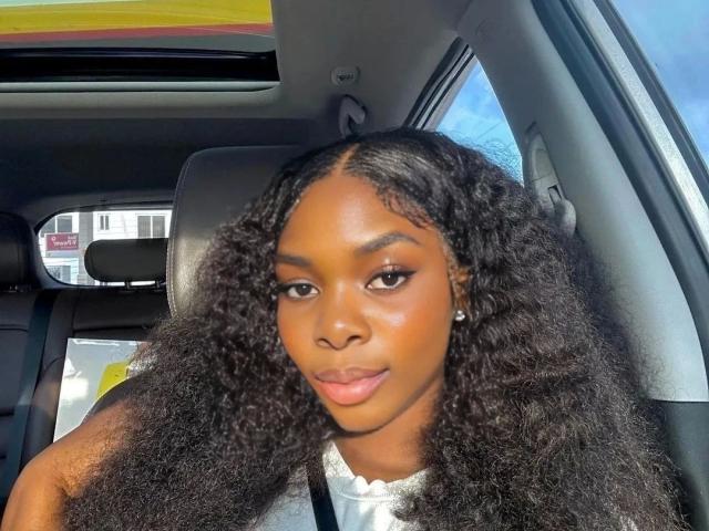 Nastybitch-hot has dark, curly hair, brown skin, and is looking at the camera with a slight smile, wearing a white top, sitting in a car.