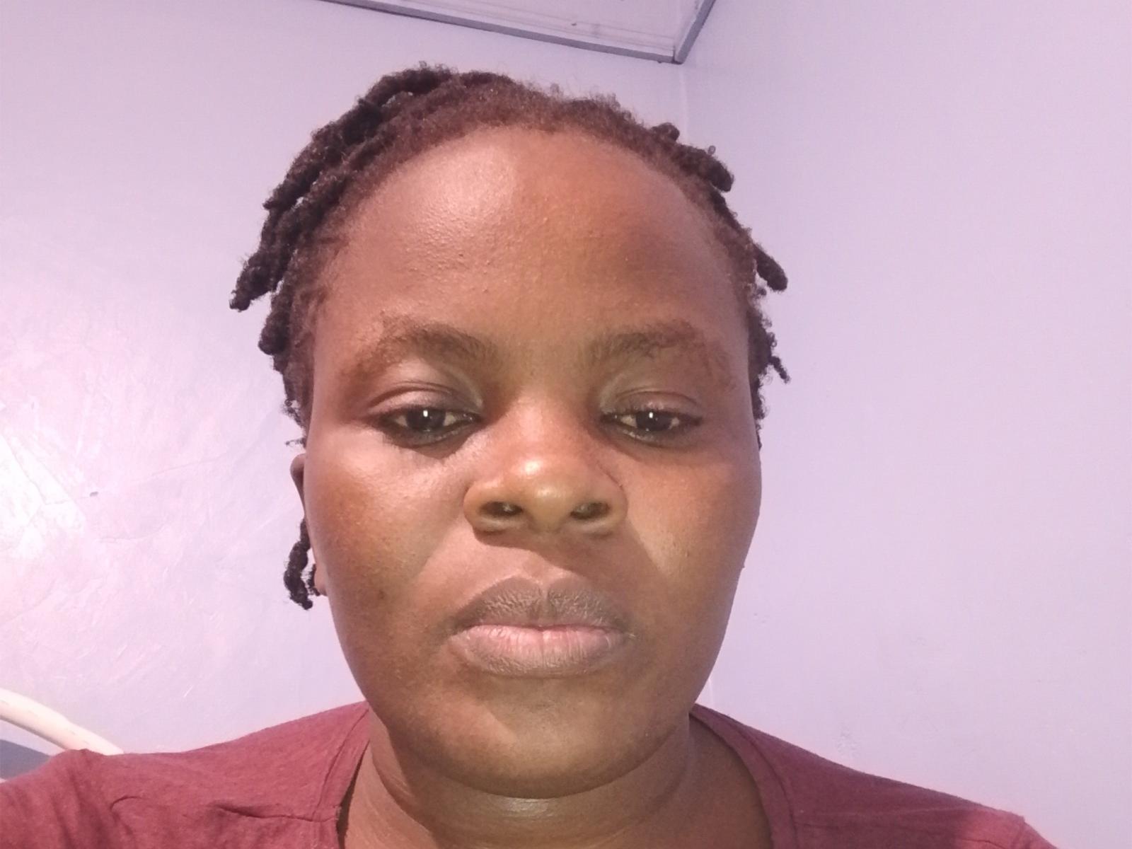PurityHot69 has dark brown skin, small nose, and short dreadlocks. She's wearing a red shirt, with a neutral expression, and slightly parted lips. Her ass is not visible.