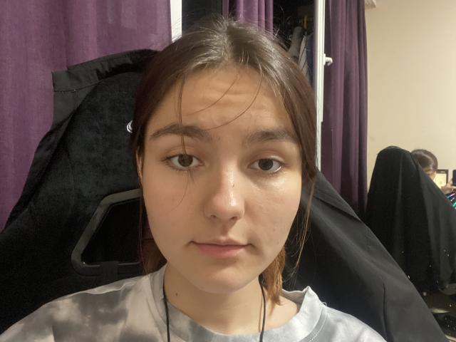 EdaPatteson tem pele clara, olhos castanhos e cabelo castanho em um estilo simples, veste uma camisa cinza, tem uma expressão ligeiramente preocupada e está usando fones de ouvido..
