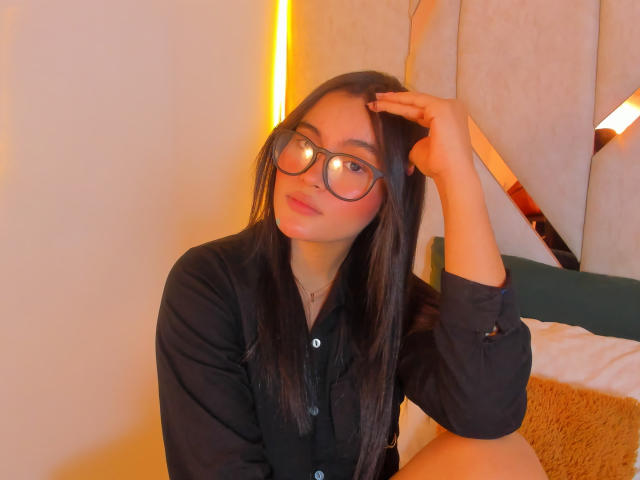 MiiaPalmer, avec ses longs cheveux foncés, ses lunettes et sa chemise noire, touchant sa tête avec une main, assise sur un lit, regardant la caméra avec un léger sourire..