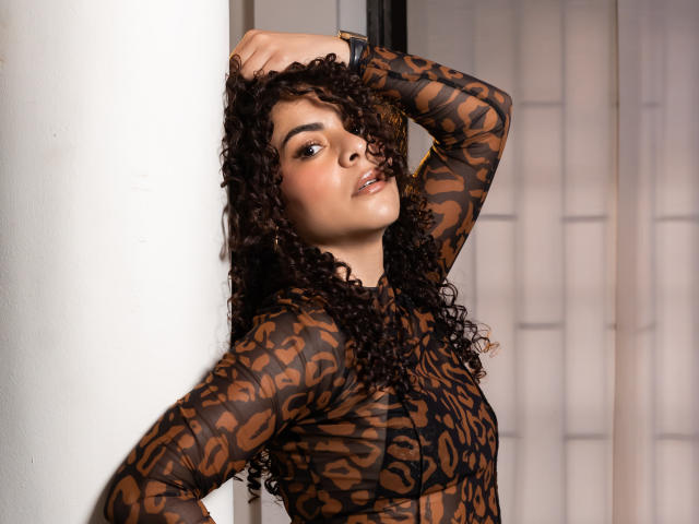 MissSunnyShine with curly brown hair, wearing a sheer leopard print top, leaning against a white wall, one arm above her head, looking at the camera with a slight smile.