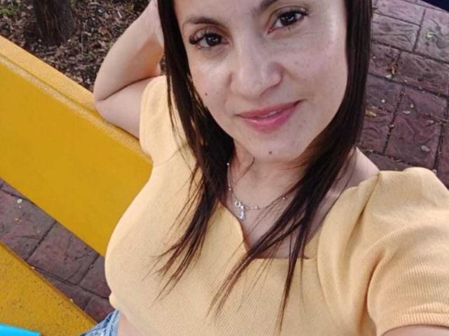 SolaraDubois with brown hair, yellow top, and silver necklace, smiling, showing small tits, hand in hair, standing by yellow and red brick.