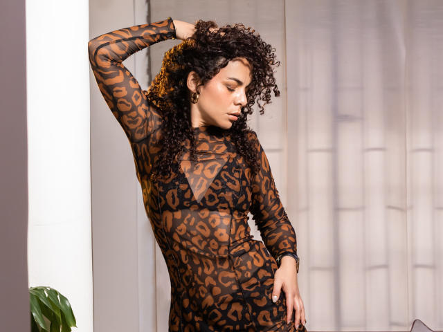 MissSunnyShine has curly brown hair, wearing a sheer leopard print dress, standing with one hand in her hair, showing her tan skin and small, perky tits.