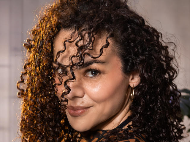 MissSunnyShine has curly brown hair, tan skin, and a cute smile. She's wearing hoop earrings and a black lace top, with her right eye slightly covered by her hair.