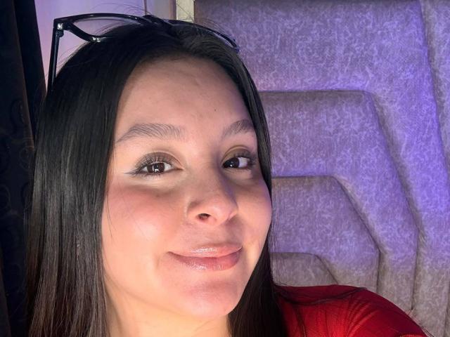 EmaJames has shiny lips, brown eyes, and straight black hair with eyeglasses on her head. She's wearing a red shirt, smiling softly. Her ass is not visible.