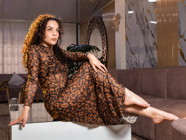 MissSunnyShine with curly brown hair, wearing a leopard-print dress, sitting on a white table, legs crossed, looking at the camera, in a modern living room.