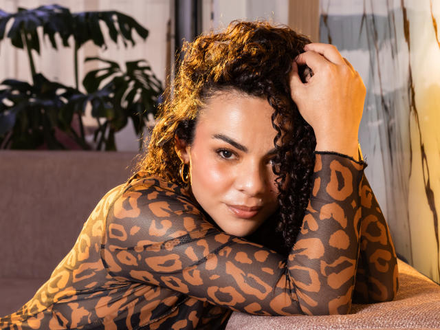 MissSunnyShine with curly brown and blonde hair, wearing a leopard print top, leans on a couch, hand in hair, smiling at the camera, showing her cute, round ass.