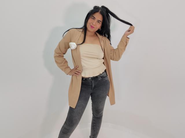 Rhouss has long black hair, pink lipstick, tan cardigan, white top, and black jeans. She's holding her hair and smiling with one hand on her hip.