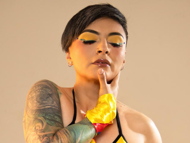 KatParadise, con cabello negro corto, sombra de ojos amarilla, un brazo tatuado y un guante amarillo, tocándose la barbilla, vistiendo un top de tirantes negro, con un fondo neutro..
