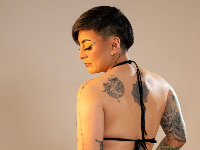 KatParadise met kort zwart haar, met de rug naar de camera, draagt een zwarte haltertop, met tatoeages op de schouders en arm, kijkt naar beneden met gesloten ogen, gouden oorbellen..